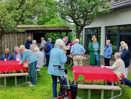 19 Peronen stehen und sitzen in einem Garten an Tischen